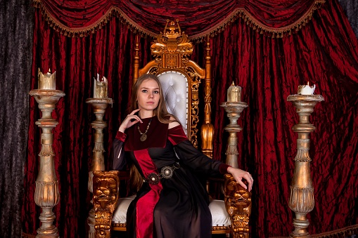Medieval queen in historical attire on golden throne in castle. Portrait of young woman in an old style dress on an antique throne in reception room of fortress. Concept of themed costume events
