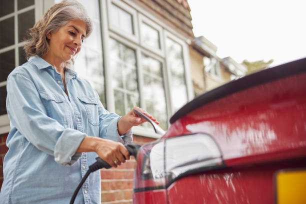 mujer madura conectando cable de carga al coche eléctrico de cero emisiones respetuoso con el medio ambiente en casa - coche doméstico fotografías e imágenes de stock