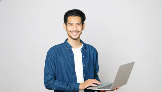 istock Young asian man holding laptop computer smiling and looking at camera standing on isolated grey background, people and technology 1326037801