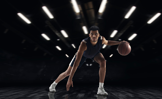 Dribbling ball. Young African sportsman, basketball player training in gym, idoors isolated on dark background with spotlights. Concept of energy, power, motion. Copyspace for ad, design. Collage, artwork