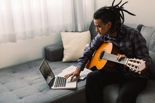 Young musician learning guitar lessons online. Young guitarist holding a guitar while watching a music tutor on his laptop. Man studying music at home during quarantine.