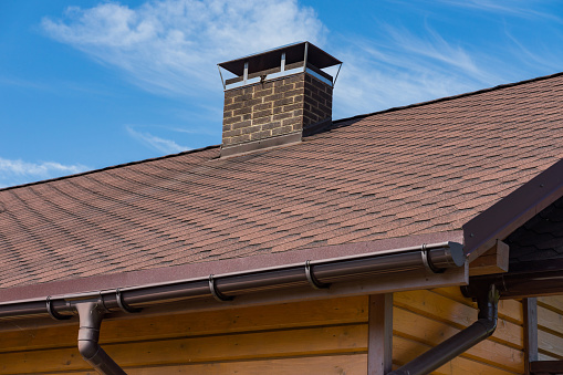 Bitumen asphalt roofing shingles and brick chimney pipe on a wooden house. Individual heating system. Rain gutter.