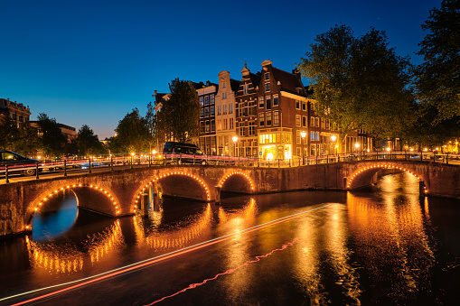 Night view of Amterdam cityscape with canal, bridge and medieval houses in the evening twilight illuminated with blurred boat light trails. Amsterdam, Netherlands
