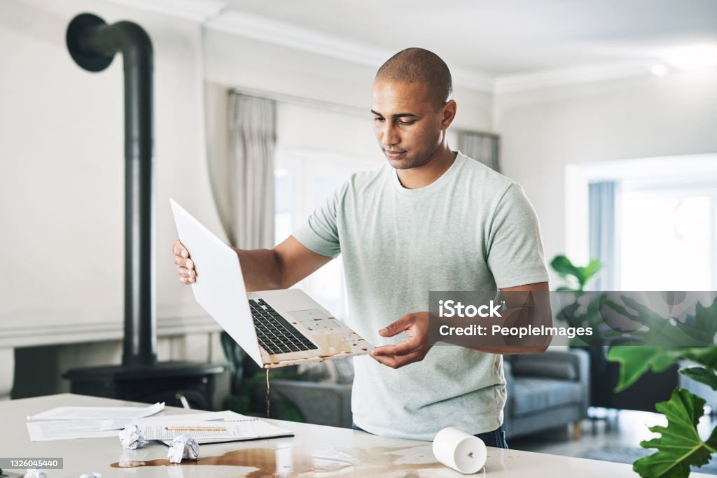 Shot of a young businessman holding a laptop damaged by coffee at home How am I going to fix this? Spilling Stock Photo