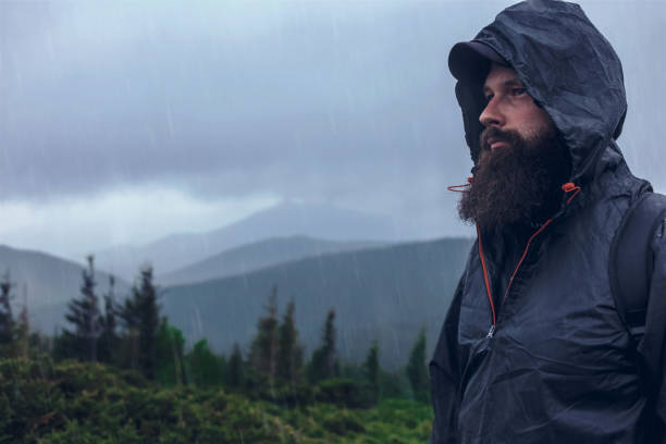 istock Handsome serious hiker bearded man on a mountain hill in a waterproof jacket under the summer rain. Mountain top against cloudy sky on natural background, copy space 1326053070