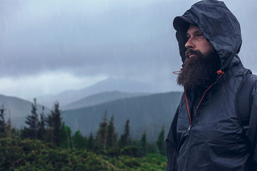 Handsome serious hiker bearded man on a mountain hill in a waterproof jacket under the summer rain. Mountain top against cloudy sky on natural background, copy space