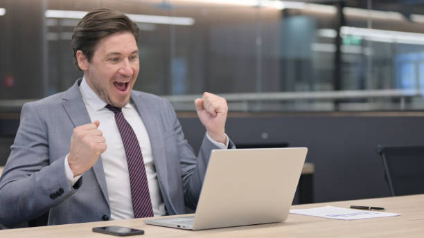 istock Middle Aged Businessman with Laptop Celebrating in Office 1326053328