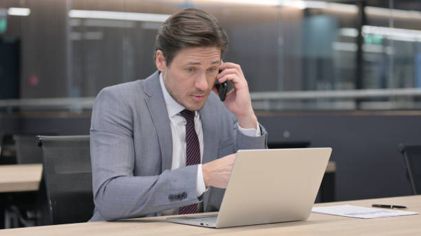 istock Angry Middle Aged Man with Laptop Talking on Smartphone 1326053501