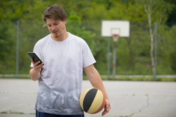 young man using mobile phone while standing on basketball court - basketball basketball hoop selective focus net imagens e fotografias de stock
