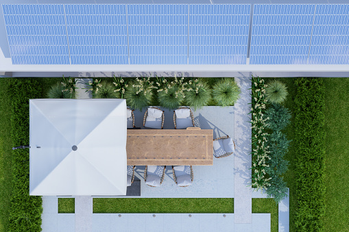 Top View Of Villa Exterior With Solar Panels On The Roof. Patio With Table, Seats And Garden With Flowers And Bushes.