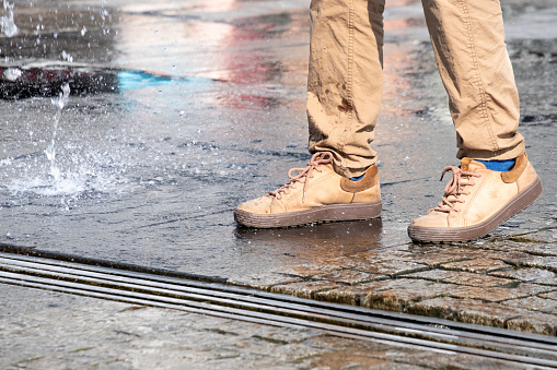 Male legs in beige pants and sneakers between the spray of a street fountain on a sunny day.