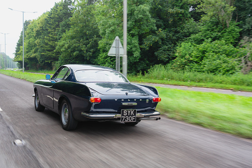 Buckinghamshire, UK - June 28, 2021: A classic dark blue Volvo 1800S from 1967 speeding down a road in the south east of England. Made famous when a white version was used by Roger Moore in the iconic sixties tv show The Saint, this svelt Swedish sports coupe is still a head-turner to this day.