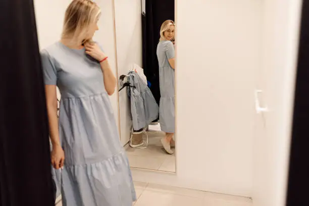 Modern young woman has opportunity to evaluate how she is wearing dress in fitting room of clothing store. Organization of sales process in supermarkets. Fashionable clothes. Shopping and pleasure. Convenience and comfort. Lifestyle.