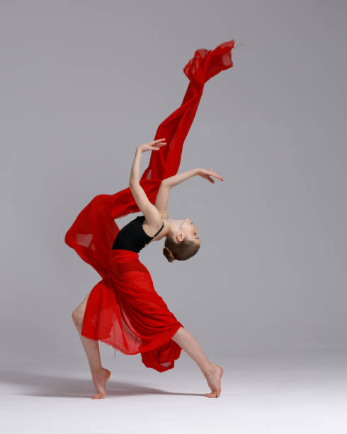 Adorable young girl in sport leotard dancing and twisting while jumping with red material against gray background