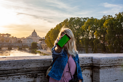 Portrait Of Young Woman Standing On Bridge Showing Cellphone With St. Peter's Basilica In Background,Italy