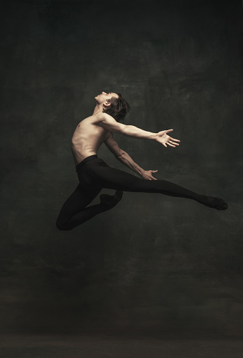 Soar above the ground. Young handsome Caucasian man, ballet dancer in art performance isolated on dark background. Art, motion, action, flexibility, inspiration concept.