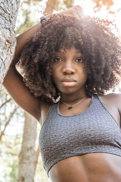 istock vertical image of young afro haired girl leaning on a tree with a gray top looking at camera and sun in the background 1326110830