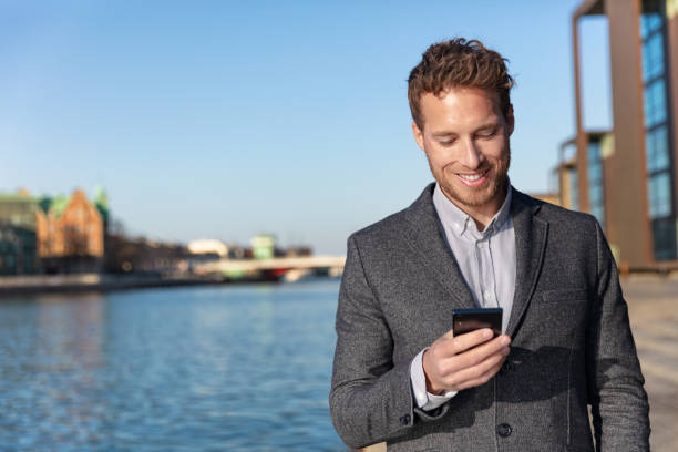 Business man texting on mobile phone using app for sms text message 5g technology. Young Caucasian businessman walking on city street outside, Copenhagen, Denmark, Europe travel stock photo