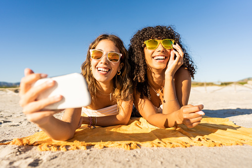 Front portrait of two cute girls with sunglasses in summer vacation using smartphone taking a selfie lying on sea sand of a tropical beach. Multiracial women travellers having fun with technology