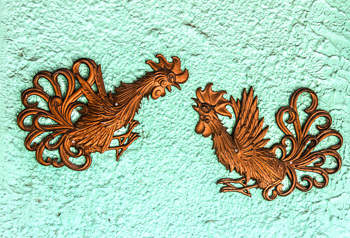 Two metal decorative fighting game roosters mounted on turquoise stucco wall swith screws