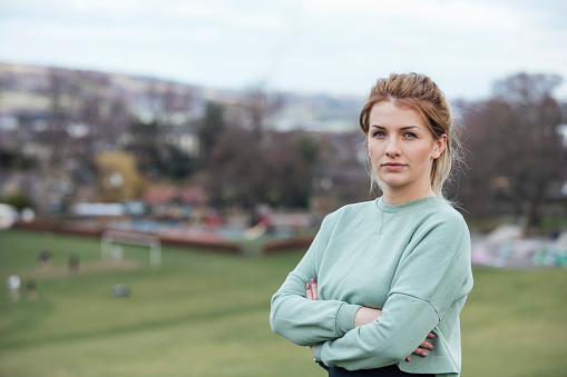 A portrait of a young caucasian woman wearing sports clothing, standing outdoors while taking a break from exercising in a public park. She is looking at the camera with her arms crossed with a serious look on her face.