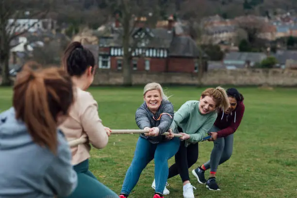 A diverse group of women exercising through a tug-of-war challenge in a public park surrounded by nature. They are there to exercise and have fun spending quality time together.