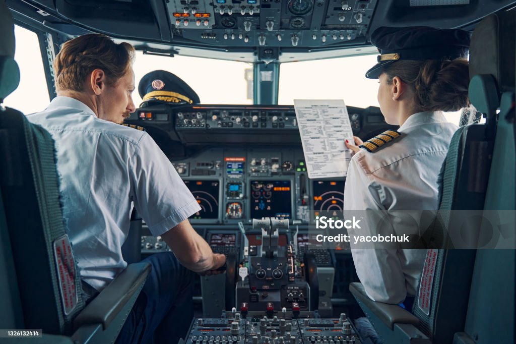 Pilot and female first officer seated in the flight deck Back view of a co-pilot with a pre-flight checklist in her hand sitting by an aircraft captain in the cockpit Pilot Stock Photo