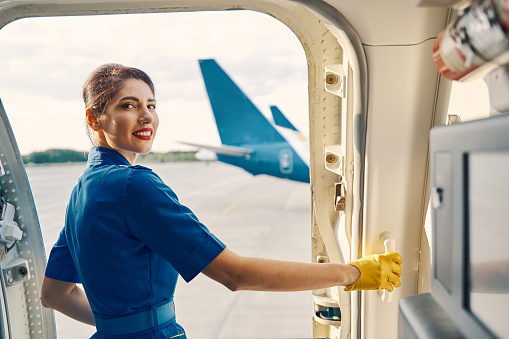 Beautiful smiling pleased dark-haired elegant flight attendant in uniform standing in the open aircraft doorway