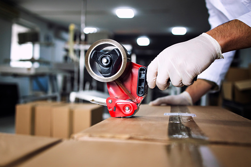 Close up view of worker's hand holding handle cutter tape dispenser and packing food in factory.