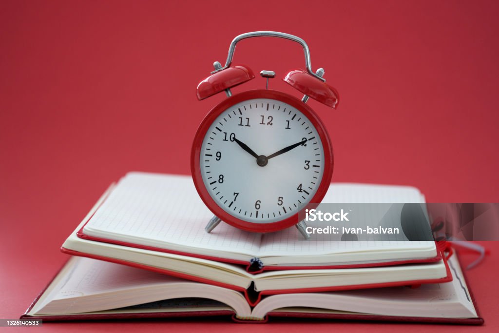 Alarm clock standing on pile of books on red background closeup Alarm clock standing on pile of books on red background closeup. Be in time by deadline concept Personal Organizer Stock Photo