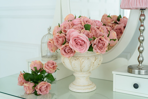 Bouquet of pink rores in vase on white boudoir table with oval mirror. Detail of the interior of the room with female boudoir