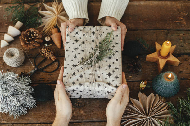 休日の装飾と素朴な木製のテーブルにスタイリッシュなクリスマスギフトトップビューを交換する手。ろうそく、糸、はさみ、松ぼっくり、モミの枝の背景に存在する家族交換 - brown pine cone seed plant ストックフォトと画像