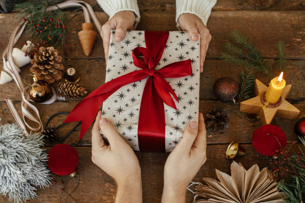 istock Hands exchanging stylish christmas gift top view on rustic wooden table with holiday decorations. Family exchanging xmas present on background of baubles, candle, pine cone, fir branches 1326147434