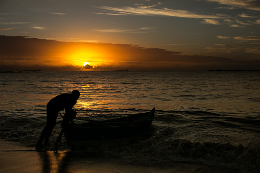 A male fisherman pushing a boat to enter the sea at sunrise against backlight.