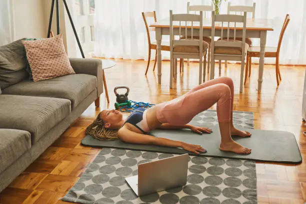 Photo of Shot of a young woman using a laptop while doing glute bridge exercises at home