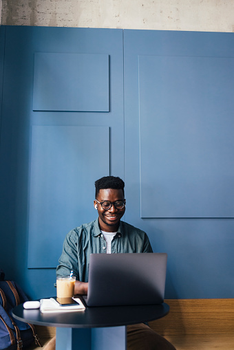 Cheerful African-American business man working in a cafe on his laptop computer and listening to music on bluetooth earphones