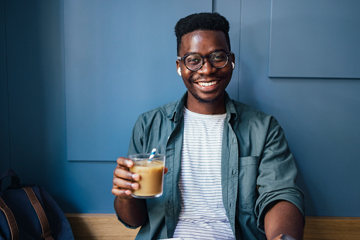 Smiling African-American business man with eyeglasses looking at camera and drinking iced coffee in a cafe while listening to music on bluetooth earphones