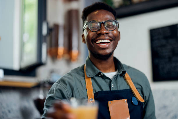 Portrait of Smiling Waiter Serving Iced Coffee in a Coffee Shop Cheerful African-American barista giving glass of iced coffee in a cafeteria barista stock pictures, royalty-free photos & images