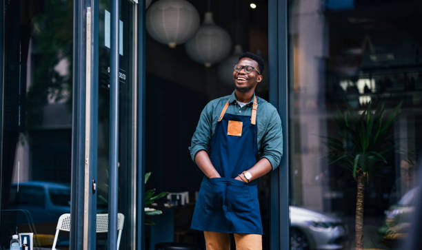 Portrait of a Confident Young Man Standing in the Doorway of his Coffee Shop Cheerful African-American barista wearing an apron and standing at the entrance of his cafe owner stock pictures, royalty-free photos & images