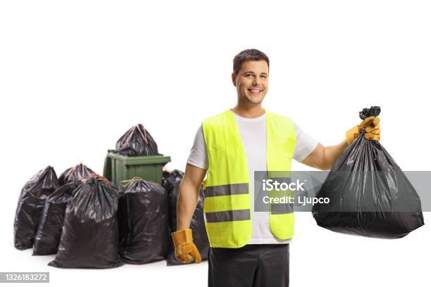 Waste Collector Holding A Plastic Bag In Front Of A Bin And A Pile Of Bags And Smiling At Camera Stock Photo - Download Image Now