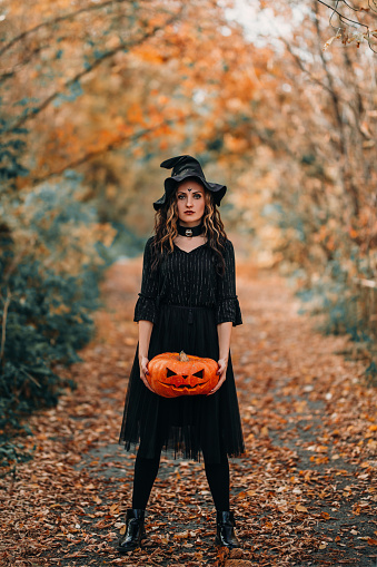 Vertical photo with girl in witch costume holding pumpkin in her hands.