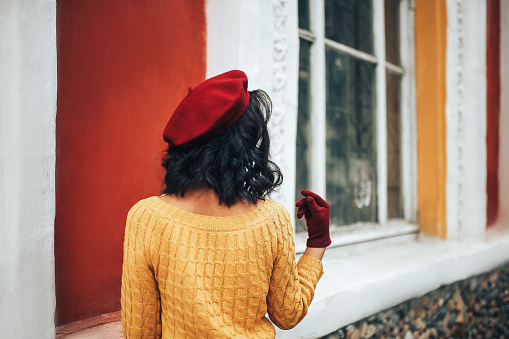 Woman in yellow sweater and red hat standing against the background of a building with large windows.