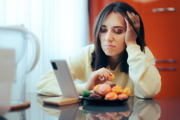 Sad Woman Stress Eating and Watching a Video on her Smartphone Unhappy person struggling with mental health and eating disorder health issues eating disorder stock pictures, royalty-free photos & images