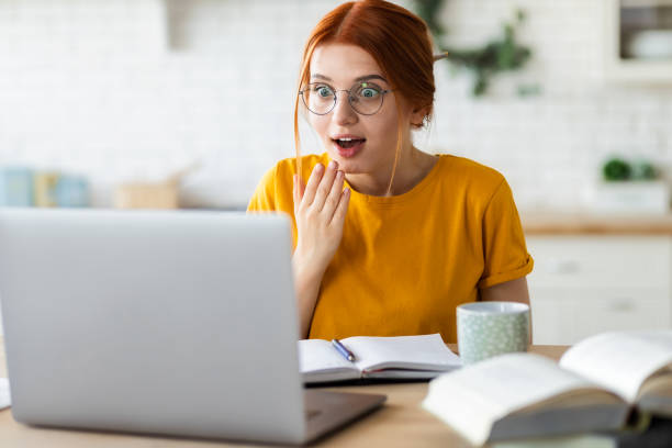 istock Surprised red-haired young girl sitting at the table with laptop computer received unexpected good news by email, emotion of surprise 1326199977
