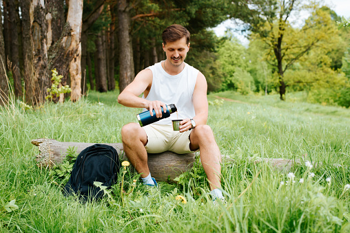 Outdoor camping. Joyful young man pouring hot drink from thermos in pine forest. Happy traveler sitting on a log and having a rest in nature during a summer hike. Active lifestyle