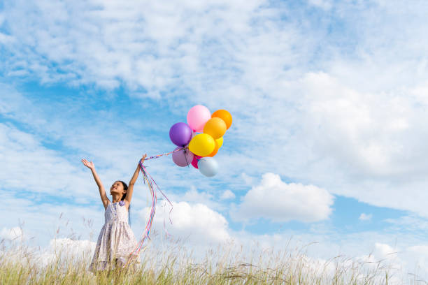 allegra ragazza carina che tiene palloncini che corrono su una nuvola bianca verde prato e cielo blu con felicità. mani che tengono vivaci mongolfiere giocano la festa di compleanno happy times estate alla luce del sole all'aperto - sun sky child balloon foto e immagini stock