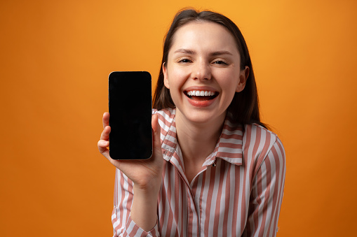 Happy smiling young woman showing you black smartphone screen with copy space against yellow background