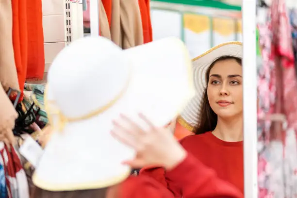 Photo of A pretty young Caucasian woman tries on a straw hat in front of a mirror in a store. Rear view, face reflected in the mirror. Shopping concept