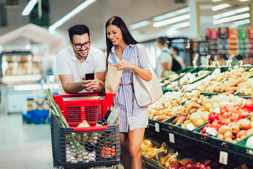 Happy couple buying vegetables at grocery store or supermarket using smart phone - shopping, food, sale, consumerism