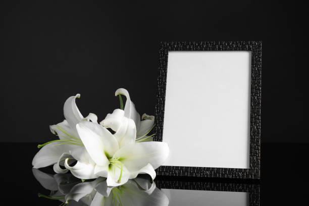 istock Funeral photo frame and white lilies on black table against dark background. Space for design 1326223088
